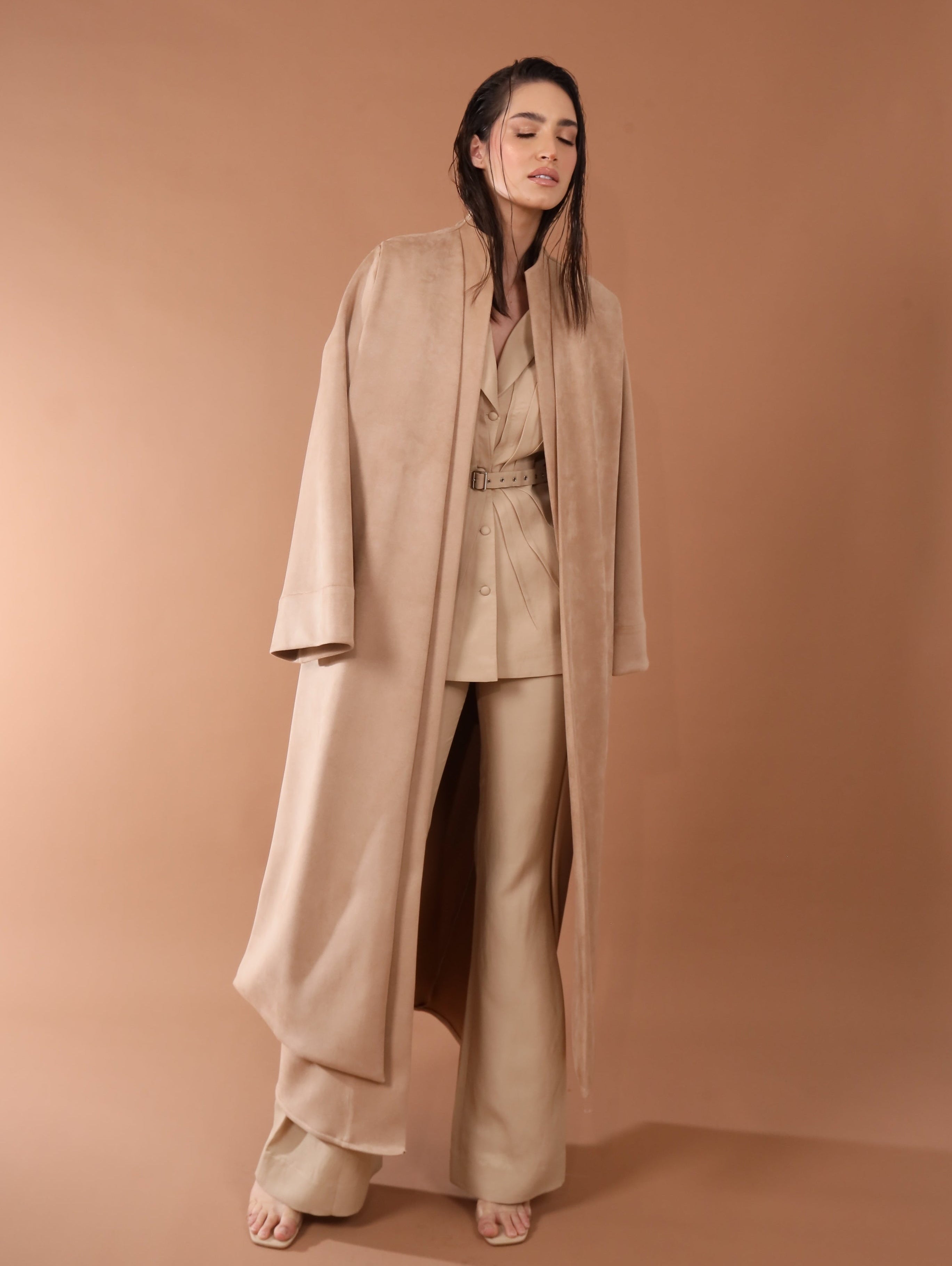 Woman wearing a beige outfit with a long coat against a brown background